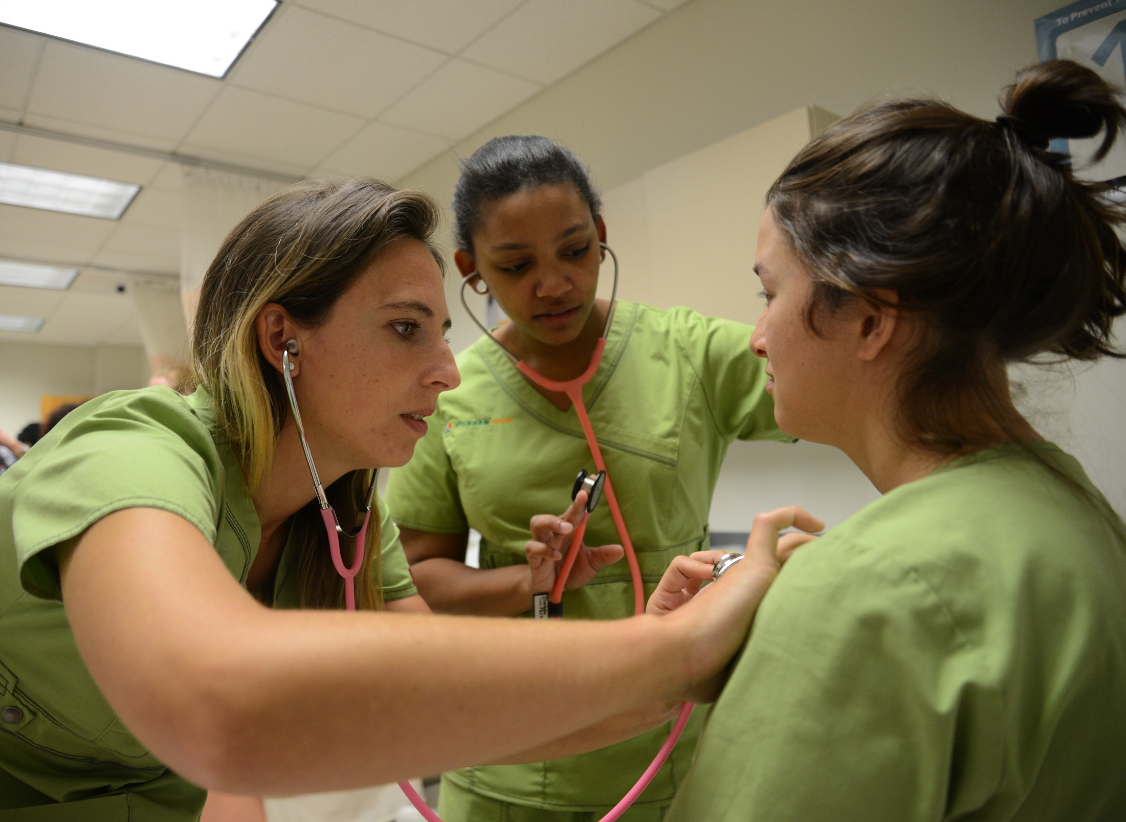 three students, one is pressing stethescope against the others chest while they sit, third student is looking on