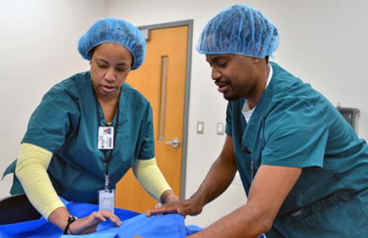 A male and female student wearing medical scrubs carefully lift a cloth sheet during a Central Sterile Processing class at Durham Tech.