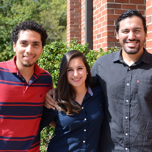 three students smiling in front of brick building also in front of bush 