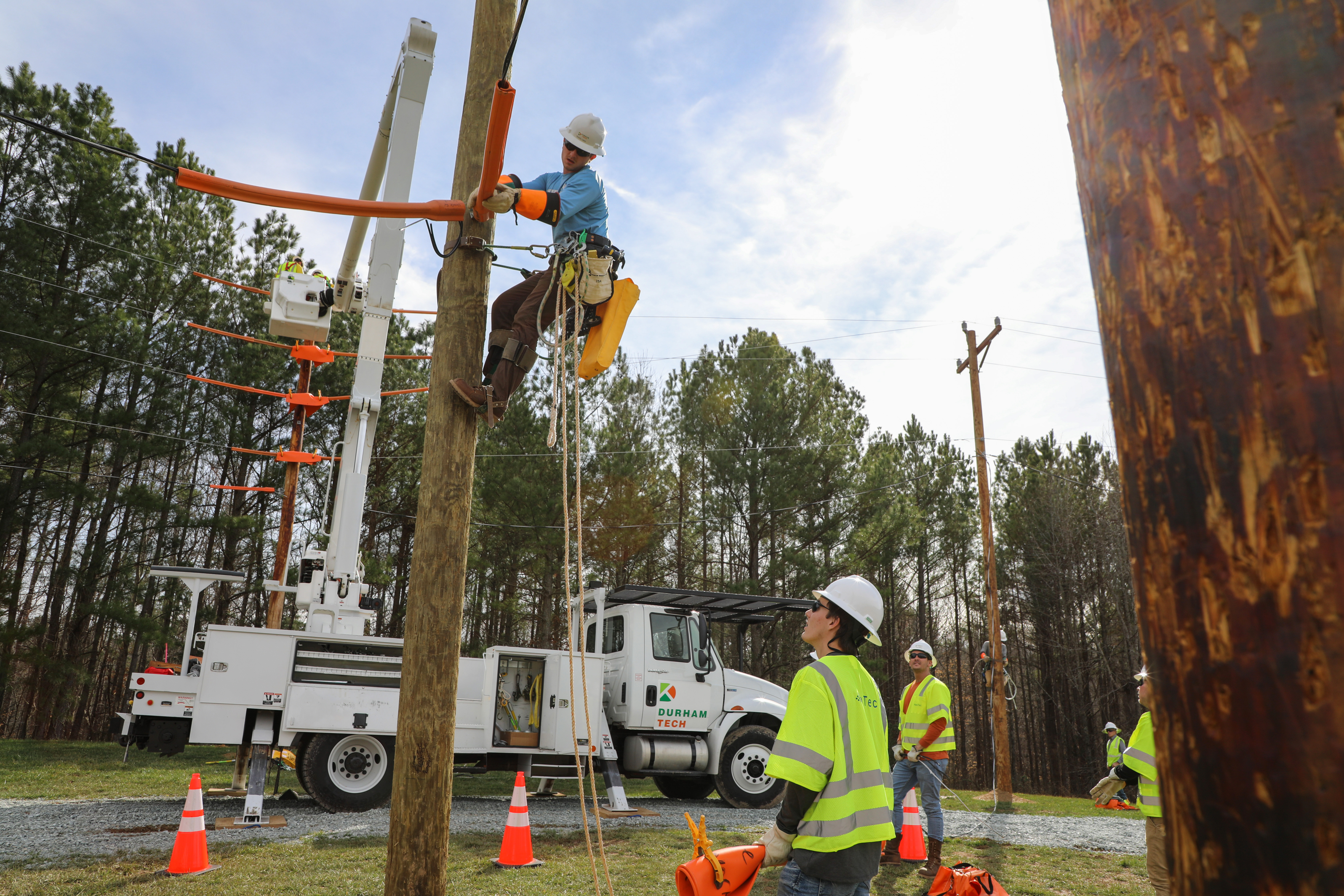 line tech students standing on ground watching another student climb a pole