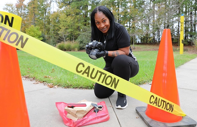 woman kneeling on side in front of evidence with camera in hand, glove on, orange cones and caution tape in front of her