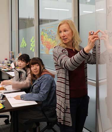 teacher standing in front of white board teaching and students sit at desks behind