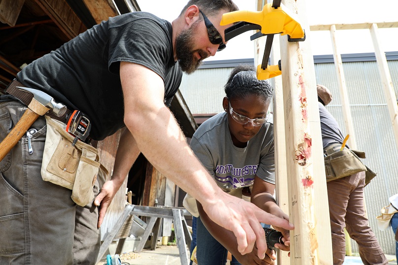 construction instructor showing student how to use construction tool, standing behind wall of shed