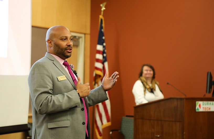 jairo standing in front of powerpoint presentation and one person in background standing at podium