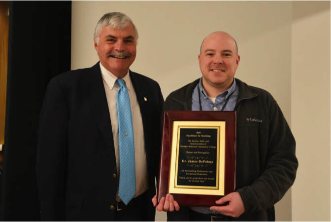 two men smiling at camera one holding an award plaque