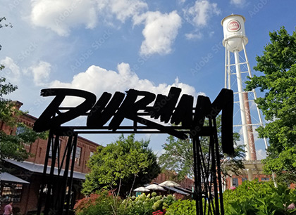 Metal sign saying Durham with the Lucky Strike logo on the water tower in the background and converted brick tobacco buildings