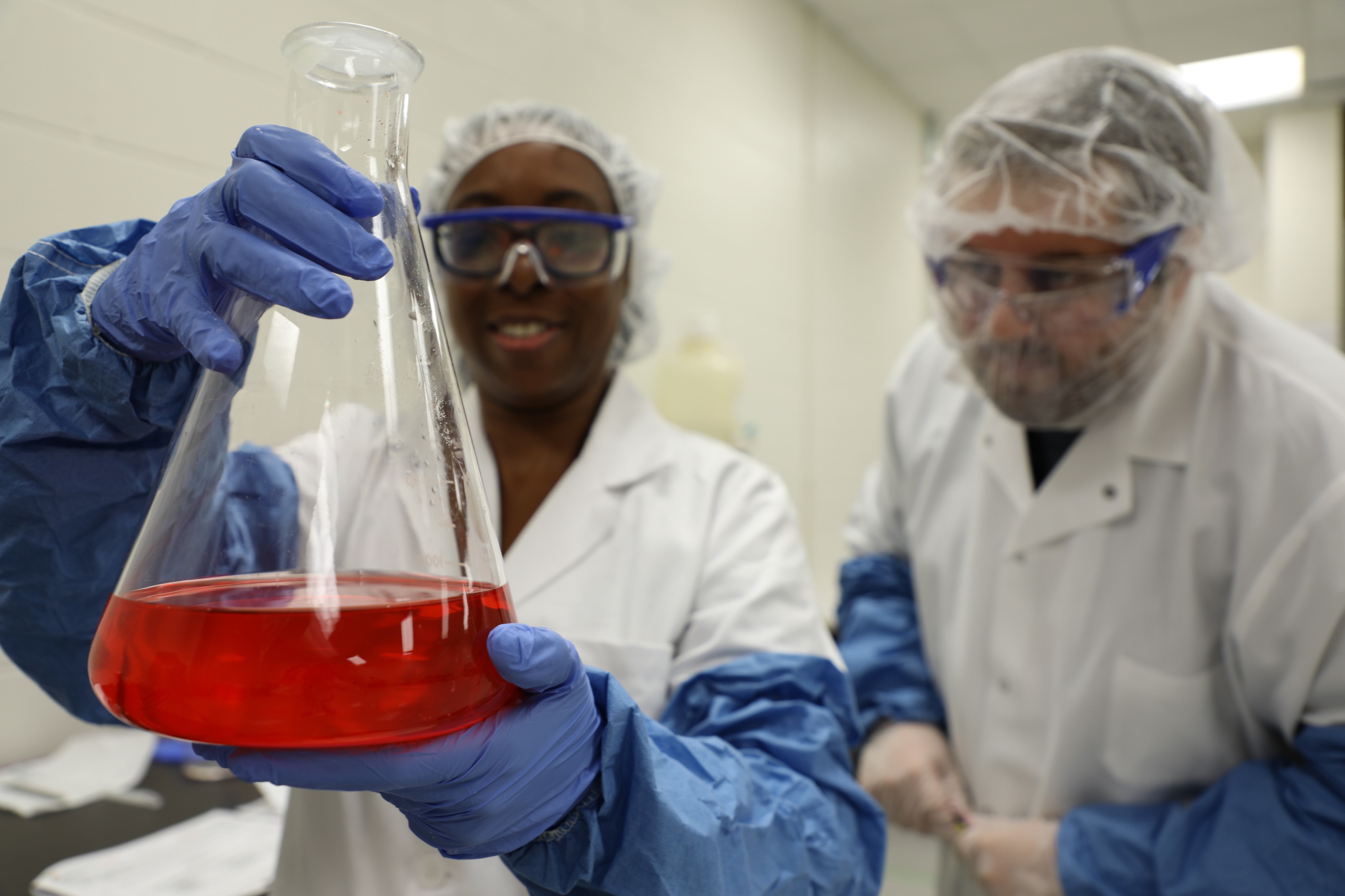 two-students-in-white-outfits-and-blue-gloves-and-goggle-holding-beaker-with-red-solution