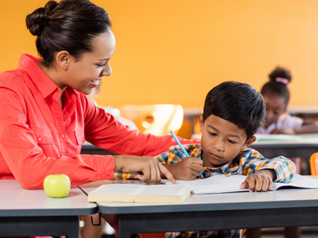 A teacher leans over and points at a book in front of a small child who is staring intently at the page with a pencil in hand.