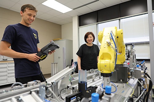 One student holds remote control for a robotic arm while another student looks on 