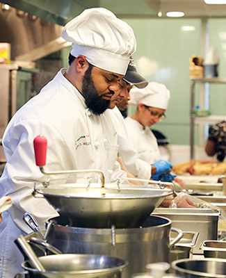 culinary student wearing white uniform works in a line of people at a table doing food prep in professional kitchen