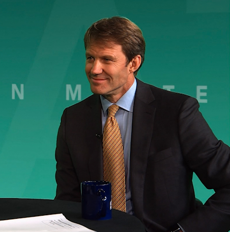 JB Buxton wearing a suit and tie sitting at a table looking to an interviewer (offscreen)