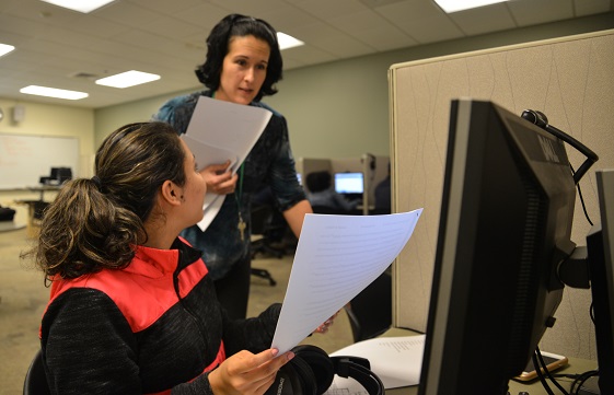 student sitting at computer and handing paper to teacher behind her