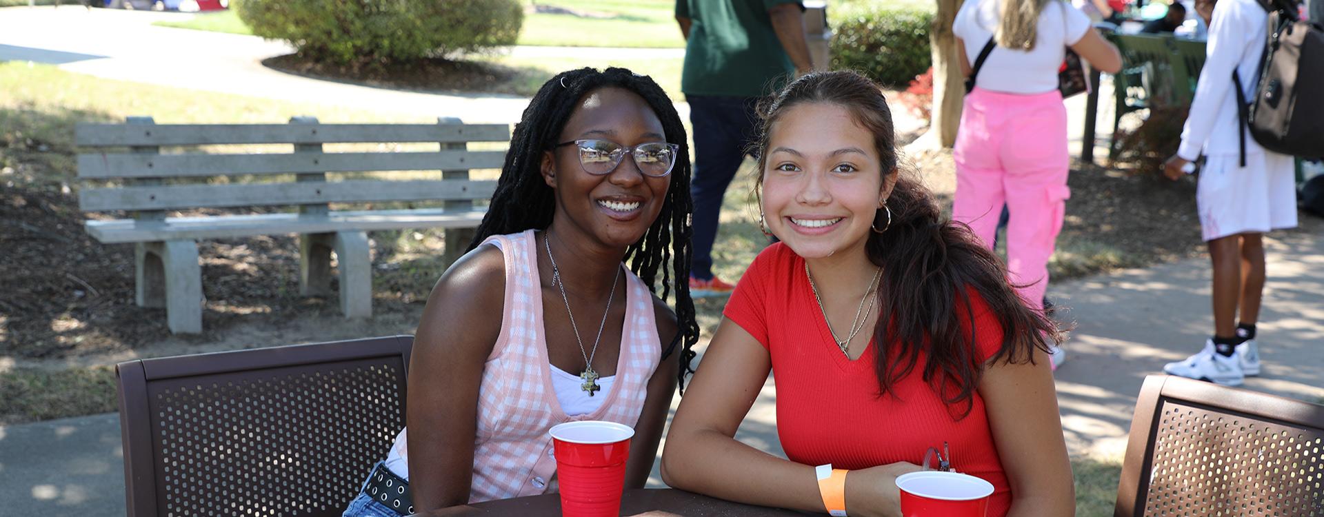 two students sitting at a table posing for the camera with students in the background