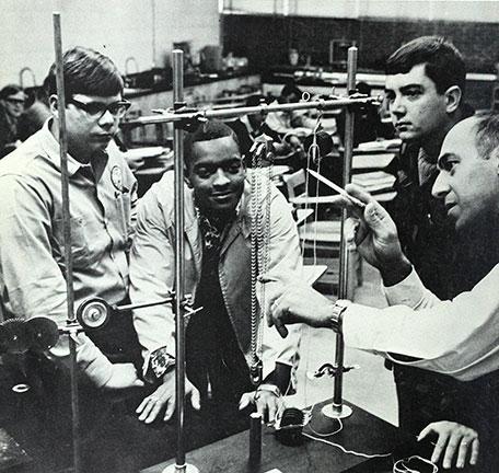 three students gather around a lab as the instructor points at a dangling chains suspended from a rod