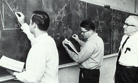 two students draw equations on blackboard as the instructor watches