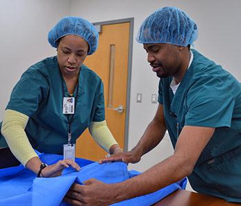 A male and female student wearing medical scrubs carefully lift a cloth sheet during a Central Sterile Processing class at Durham Tech.