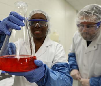 two-students-in-white-outfits-and-blue-gloves-and-goggle-holding-beaker-with-red-solution