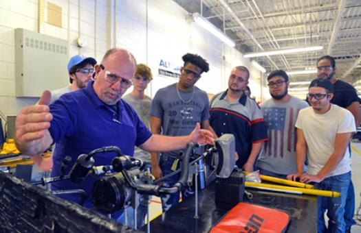 An instructor in the Automotive Systems Technology program uses right hand to indicate length for a for a group of eight Durham Tech students.