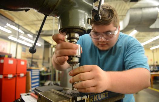 machining student works on milling machine, leaning over looking closely and wearing goggles