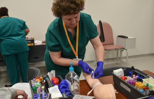student wearing purple gloves and practicing putting needle in arm of mannequin