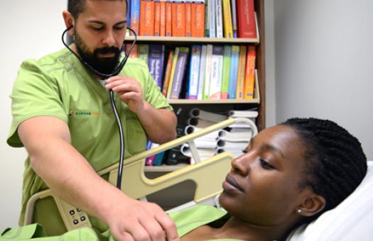 A male student wearing medical scrubs practices using a stethoscope on a female student while she lays on a gurney.