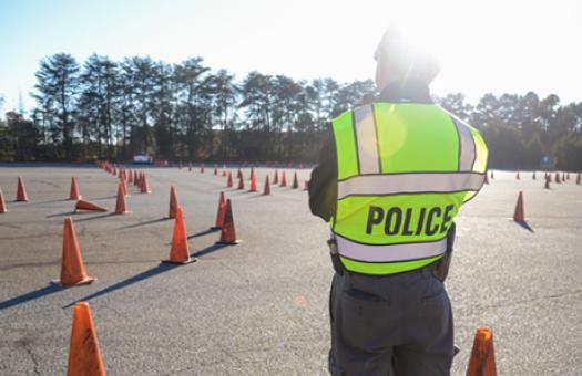 A Basic Law Enforcement Training instructor stands in uniform with a neon yellow police vest on while looking out over a road training course with orange cones.