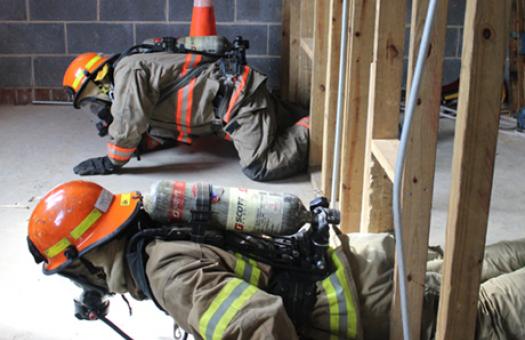 Two student firefighters crawl from right to left through a gap between wooden planks during a training exercise.