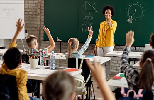 An image of an elementary school teacher choosing a student from a classroom full of students with raised hands