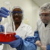 two-students-in-white-outfits-and-blue-gloves-and-goggle-holding-beaker-with-red-solution