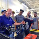 An instructor in the Automotive Systems Technology program uses right hand to indicate length for a for a group of eight Durham Tech students.