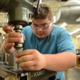 machining student works on milling machine, leaning over looking closely and wearing goggles