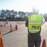 A Basic Law Enforcement Training instructor stands in uniform with a neon yellow police vest on while looking out over a road training course with orange cones.