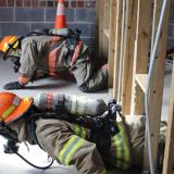 Two student firefighters crawl from right to left through a gap between wooden planks during a training exercise.